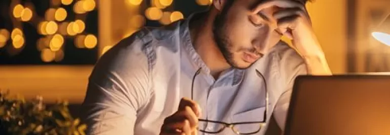 Persona sentada frente a una computadora, sosteniendo gafas, con luces brillantes de fondo.