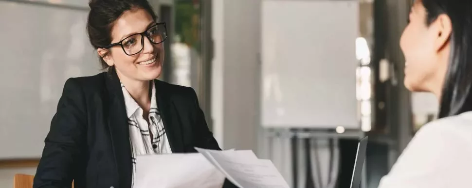 Dos personas conversando en una oficina, una sostiene documentos.