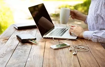 Persona trabajando en un portátil al aire libre, sosteniendo una taza de café.